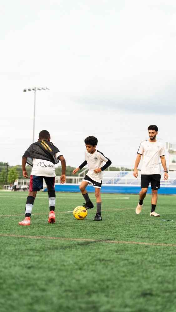 A player dribbling the ball past a defender while a coach looks on